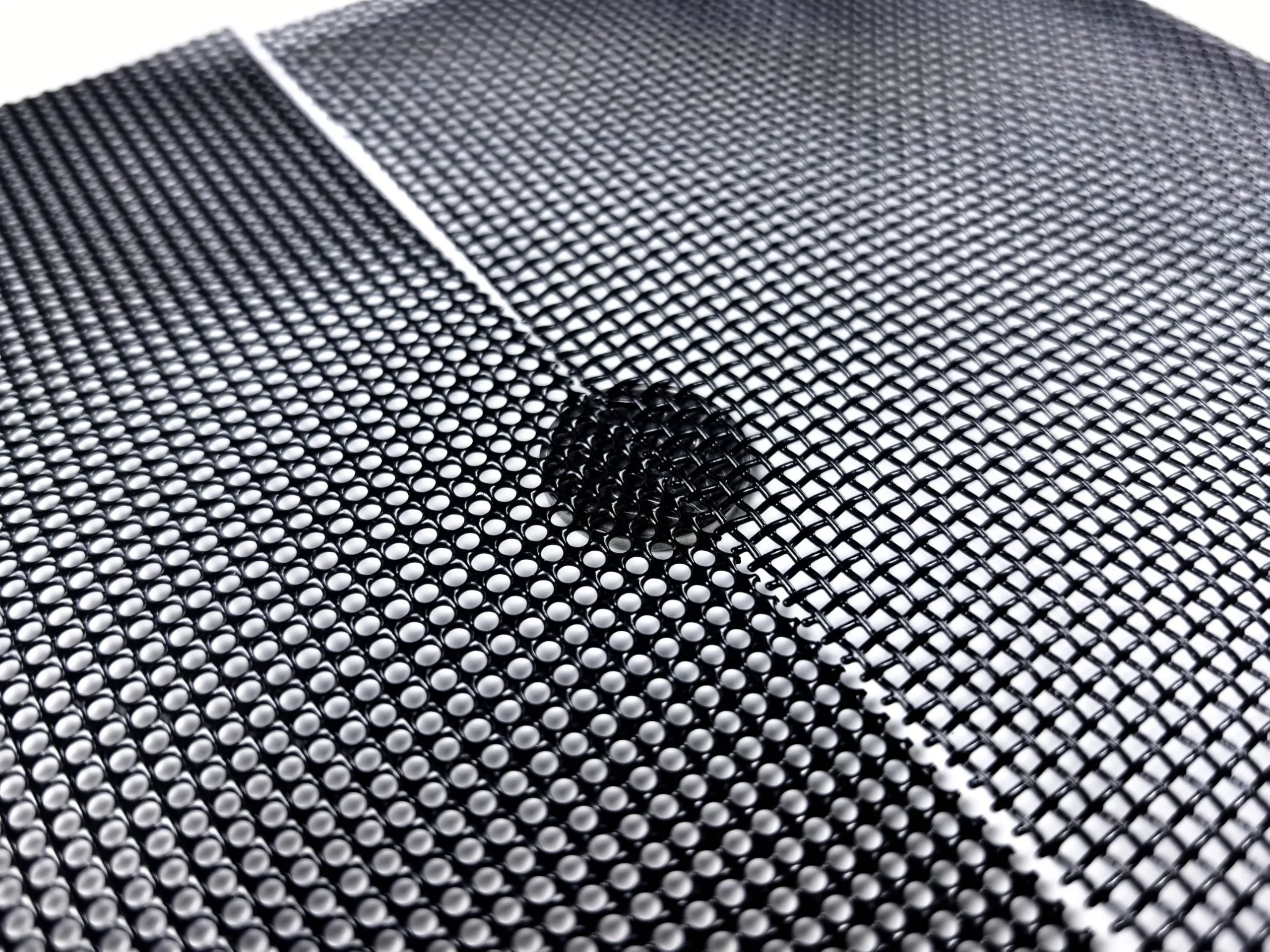 Factory photo of black powder-coated security screen mesh: round-hole finish on the left and square-hole finish on the right, with a small disc of square-hole mesh placed on the round-hole sheet